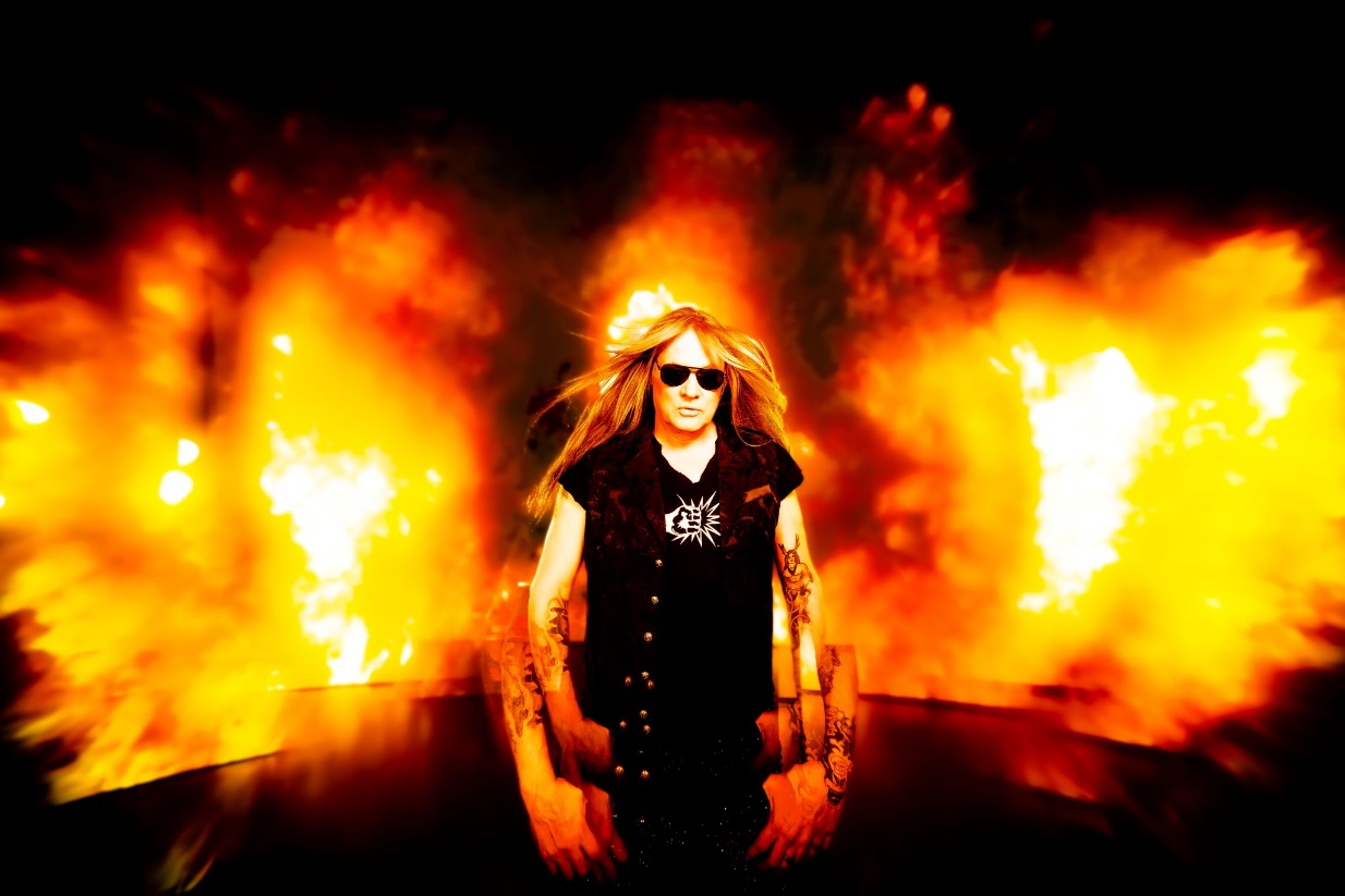 A person with long hair wearing sunglasses and a black vest with a fire behind him

Description automatically generated