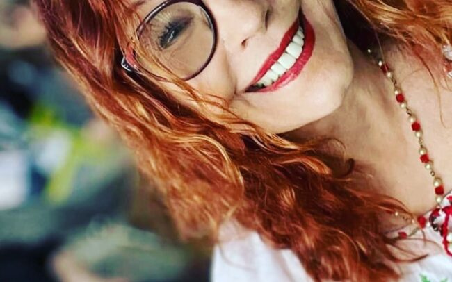 A person with red hair and glasses smiling Description automatically generated